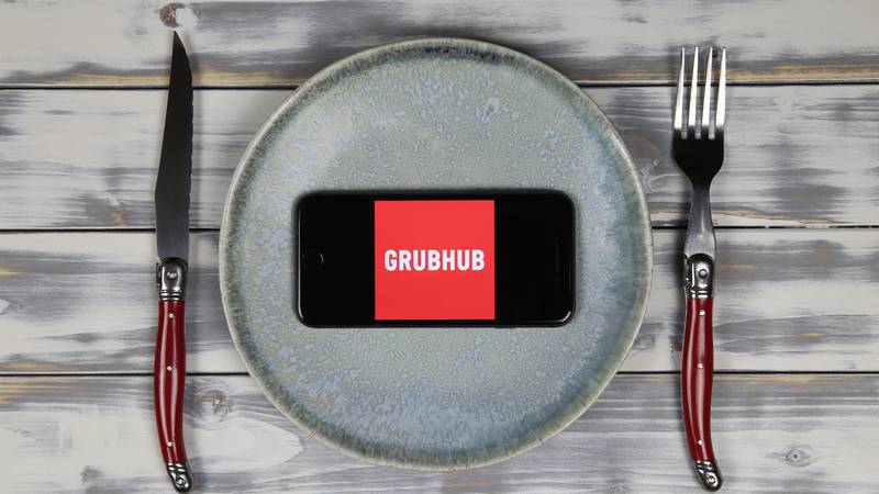 Close up of mobile phone screen with logo lettering of food delivery service grubhub on wood table with dish and cutlery (focus on center)